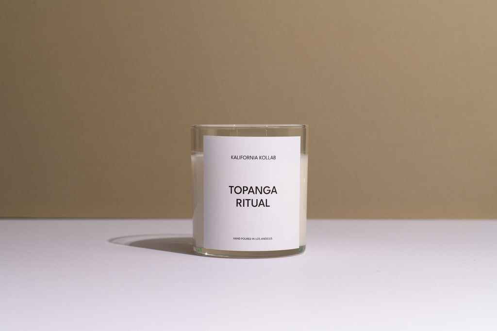 A clear glass jar candle with white wax and a white label reading 'CALIFORNIA COLLAR' and 'TOPANGA RITUAL', set on a white surface against a warm beige background.