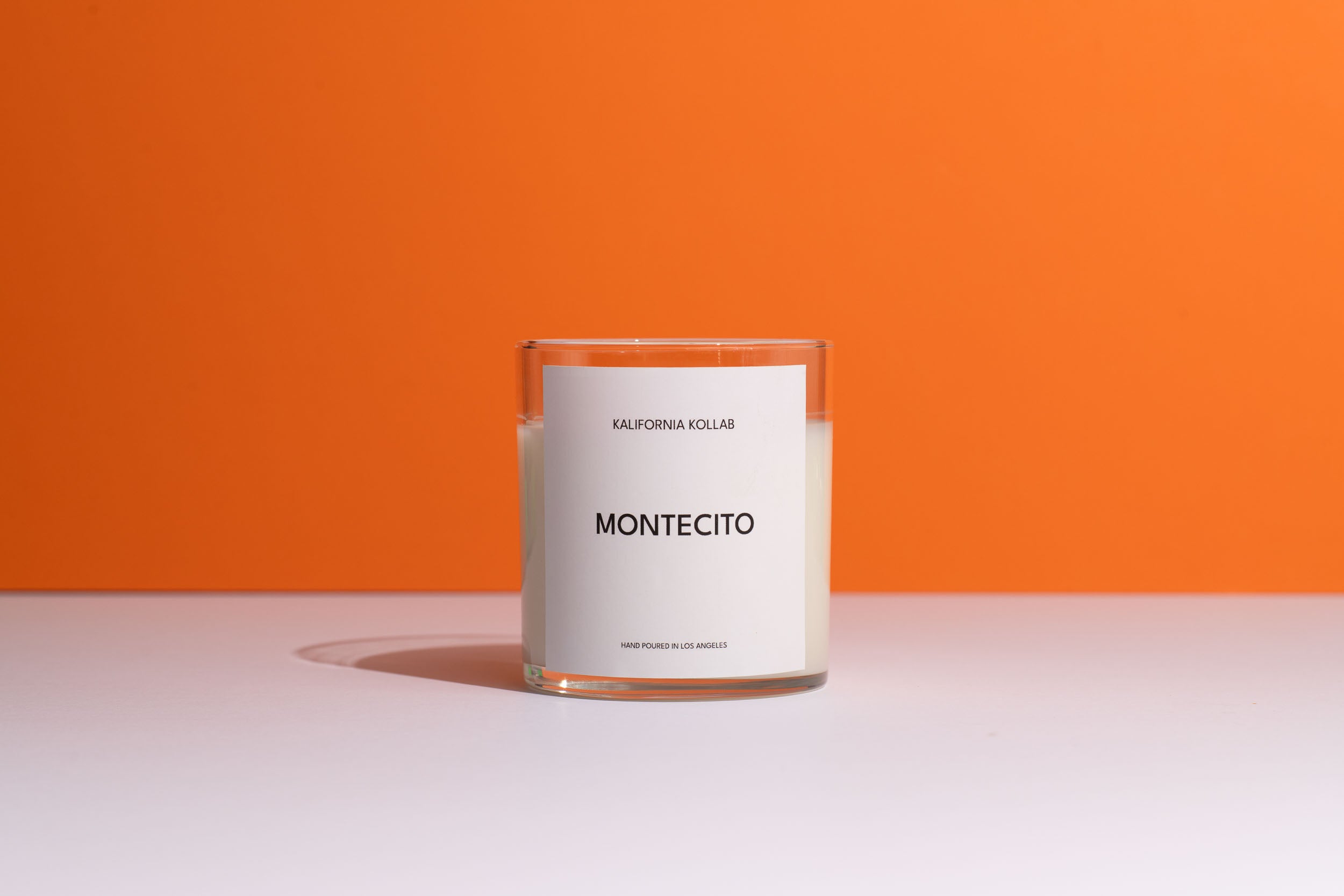 A glass jar candle with white wax and a minimalist white label reading 'KALIFORNIA KOLLAB' and 'MONTECITO', set against a solid orange background and white surface.