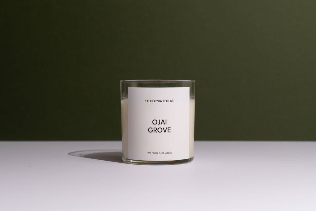A clear glass jar candle with white wax and a minimalist white label reading 'OJAI GROVE' and 'CALIFORNIA COLLEGE', set on a white surface against a dark olive green background.