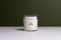 A clear glass jar candle with white wax and a minimalist white label reading 'OJAI GROVE' and 'CALIFORNIA COLLEGE', set on a white surface against a dark olive green background.