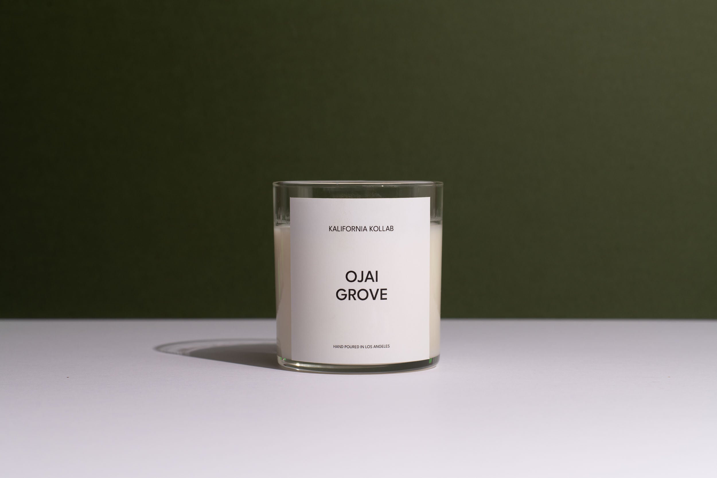 A clear glass jar candle with white wax and a minimalist white label reading 'OJAI GROVE' and 'CALIFORNIA COLLEGE', set on a white surface against a dark olive green background.