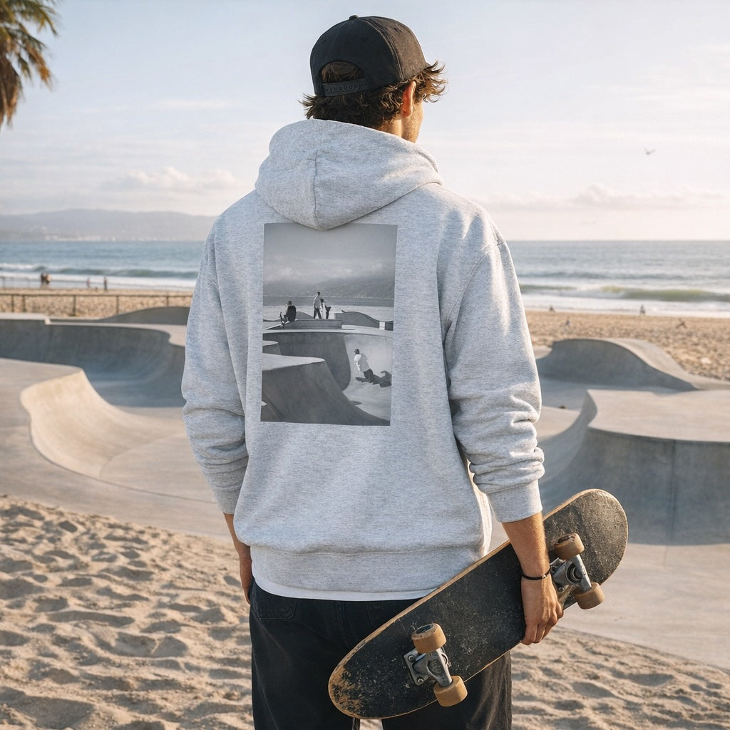 Person wearing a gray hoodie with a skateboarder graphic, standing at a skate park with ocean view.